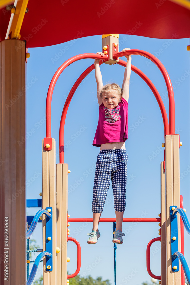 A little active girl spends time on a walk on the playground in the yard, hanging in her arms on iron arcs. 