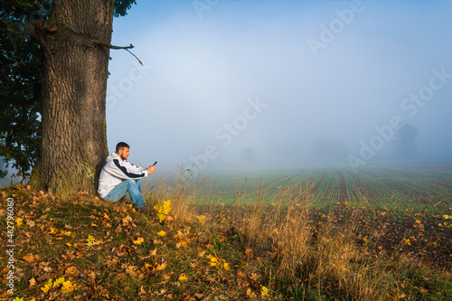Young caucasian man from side sitting under large autumn colorful foliage tree using his mobile phone, relax at sunrise with misty fog. Czech landscape, copy space
