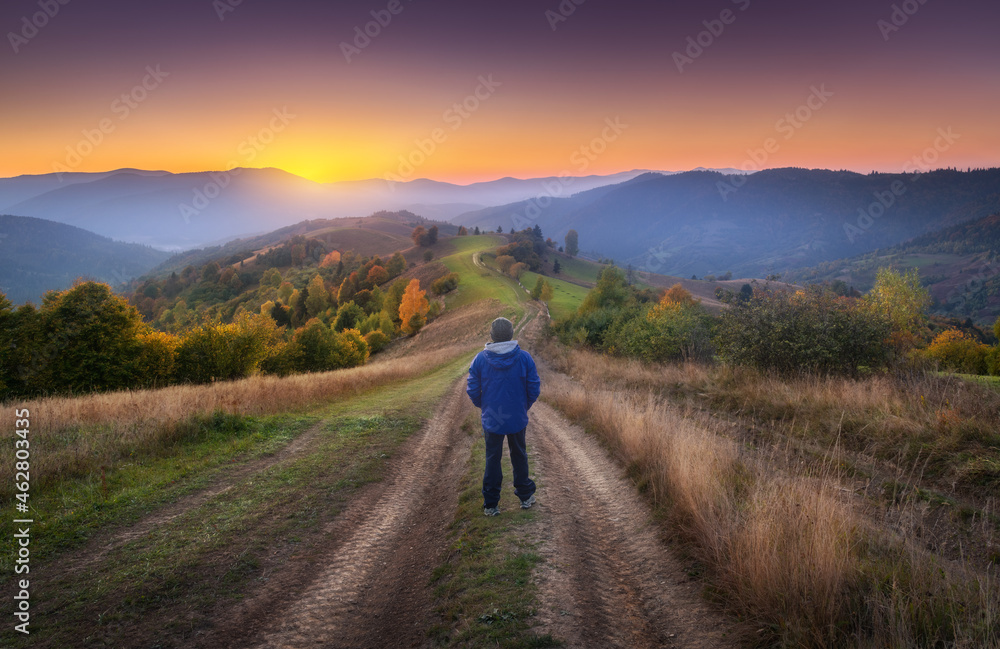 Man on the rural dirt road on the hill looking on mountains in fog at colorful sunset in autumn in Ukraine. Landscape with guy, foggy hills, green grass, meadows, trail, forest, orange sky in fall