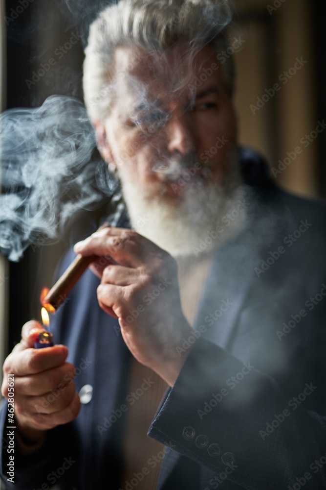Fototapeta premium Portrait of handsome grey haired mature man lighting up cigar while standing indoors