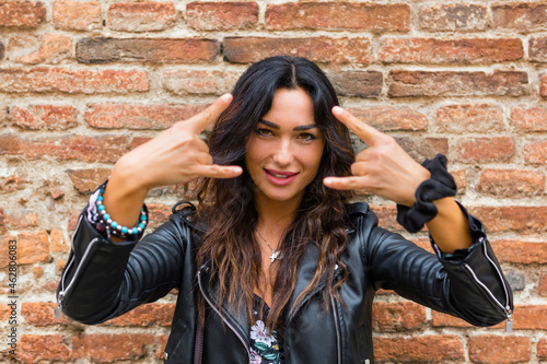 Portrait of young woman wearing black leather jacket and showing rock and roll sign, brick wall