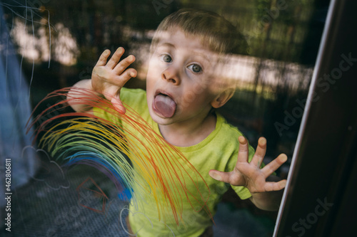 Portrait of little boy standing behind balcony door licking glass pane with drawn rainbow photo