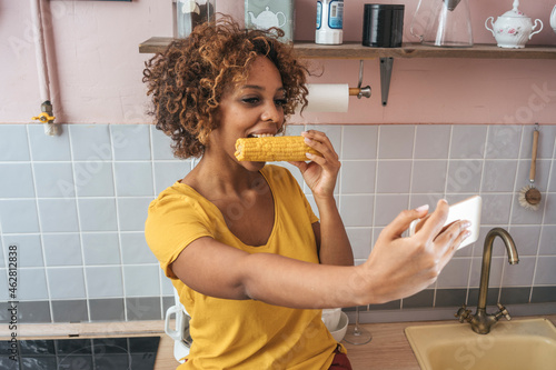 Young woman eating a corncob and taking a selfie in kitchen