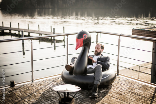Hip businessman working on a houseboat, sitting a floating swan, using digital tablet photo