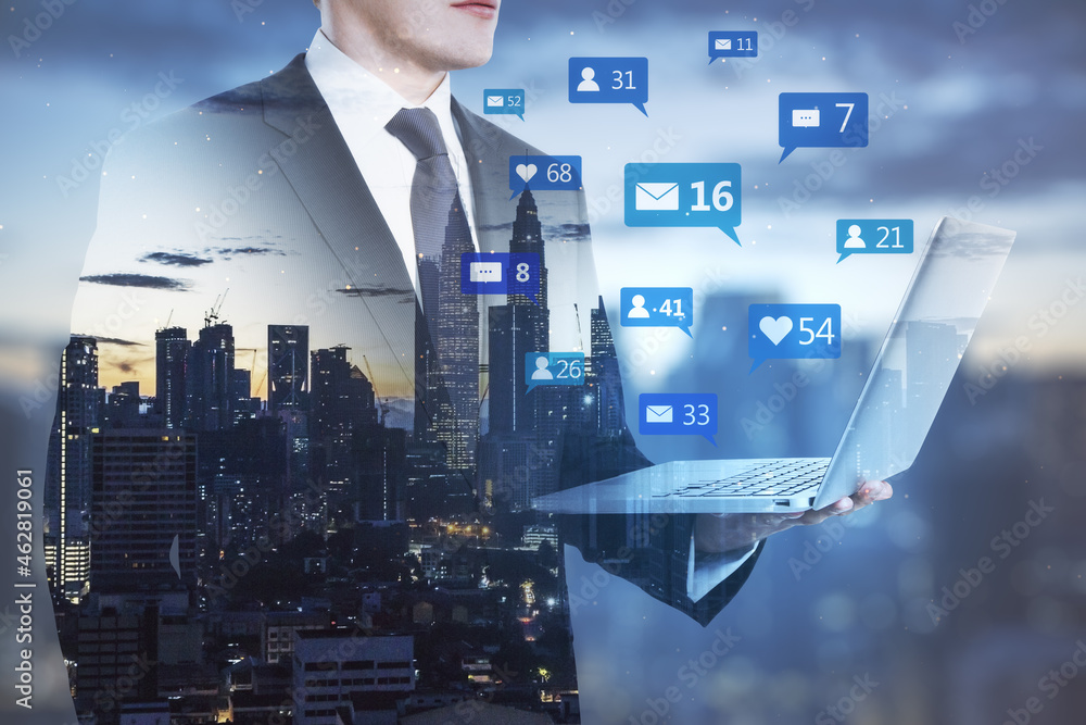 Obraz premium Businessman holding laptop with social media icons on blurry night city background. Communication, network, connections, device and app concept. Double exposure.