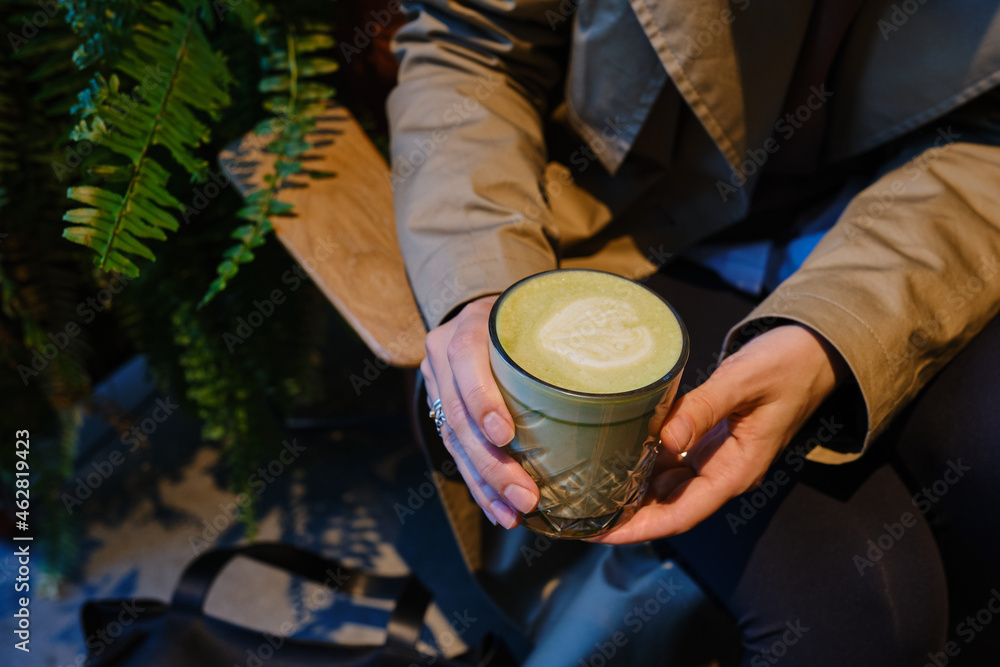 Woman with a cup of matcha latte sits in a cozy cafe. Delicious green matcha tea in a transparent glass close-up. Japanese hot milky beverage. Healthy Matcha Tea Made with Organic Ingredients