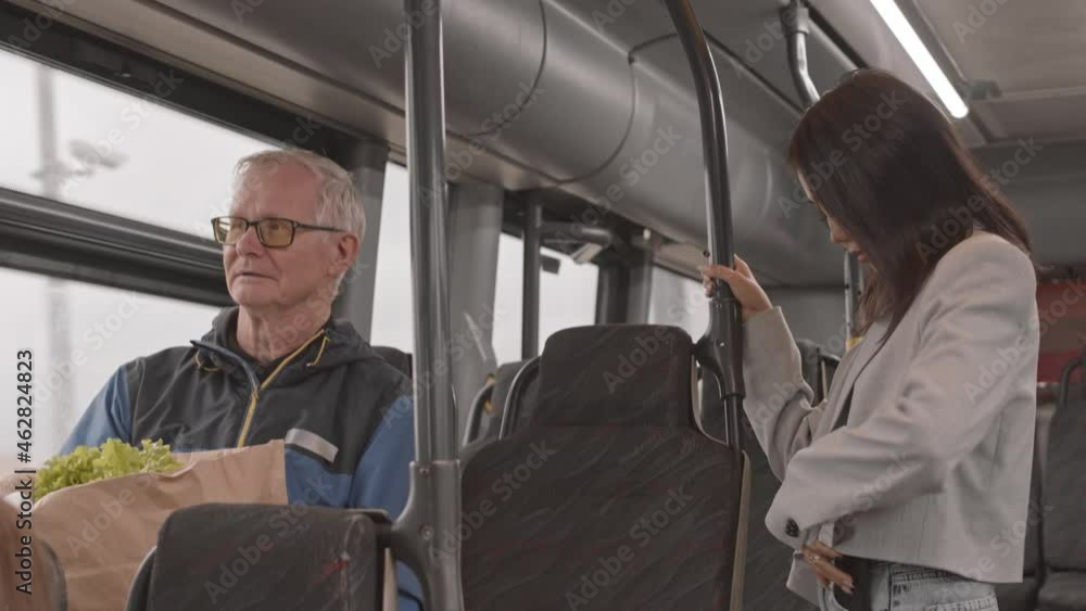 Medium of young Asian woman standing in bus, holding handrail, senior Caucasian man holding paper bag with groceries sitting on seat by window Medium of young Asian woman standing in bus, holding handrail, senior Caucasian man holding paper bag with groceries sitting on seat by window