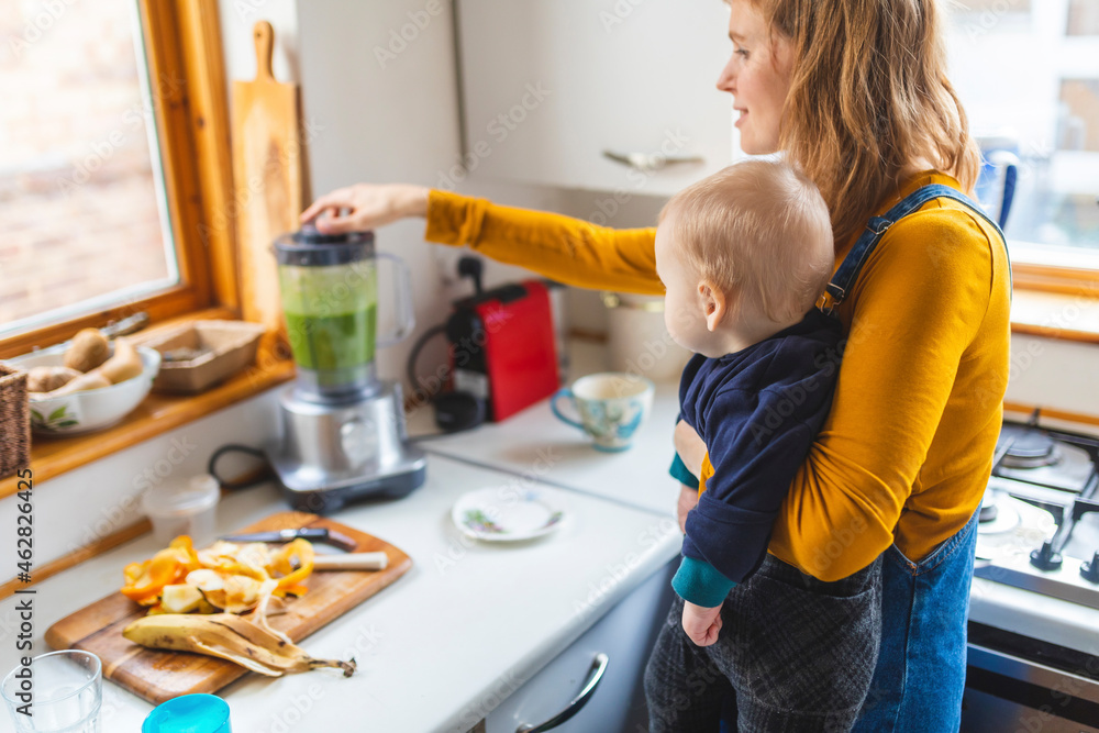 Woman in the kitchen preparing healthy smoothie and holding her baby son Woman in the kitchen preparing healthy smoothie and holding her baby son