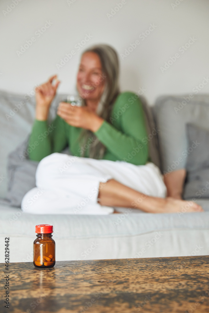 Smiling woman showing pill with drinking water on sofa at home Smiling woman showing pill with drinking water on sofa at home