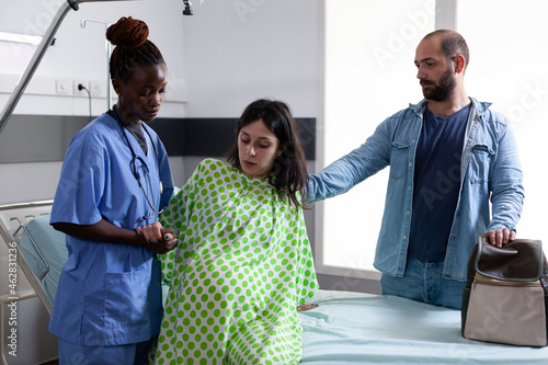 Black obstetrics nurse helping pregnant woman get out of bed in hospital ward at facility. Person with pregnancy belly receiving support from african american assistant and father of child photo