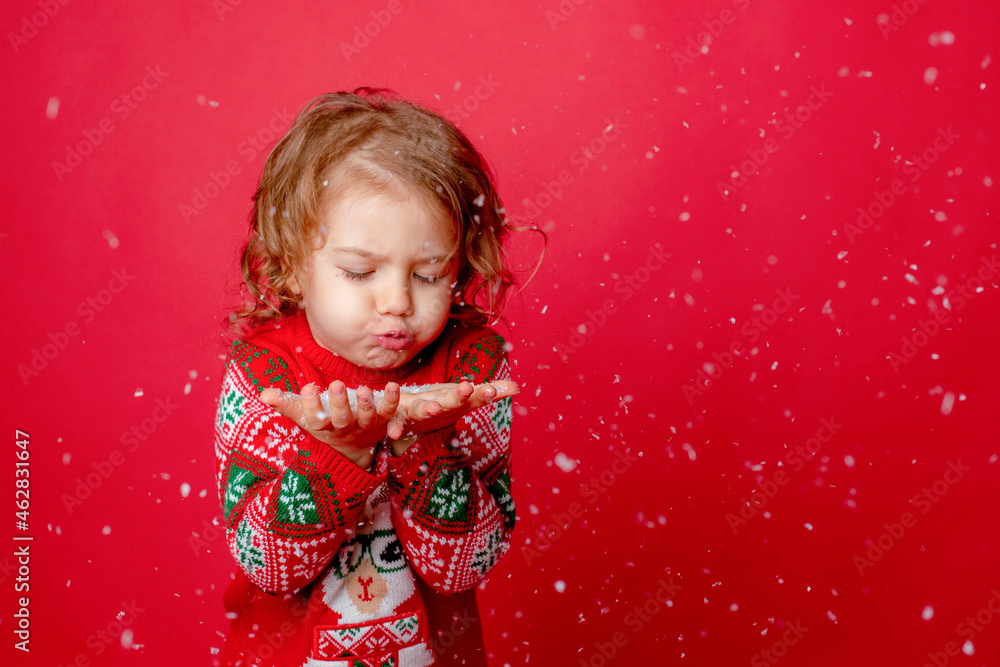 Cute little girl in a red sweater on a red background blowing snow, Christmas, New Year Cute little girl in a red sweater on a red background blowing snow, Christmas, New Year