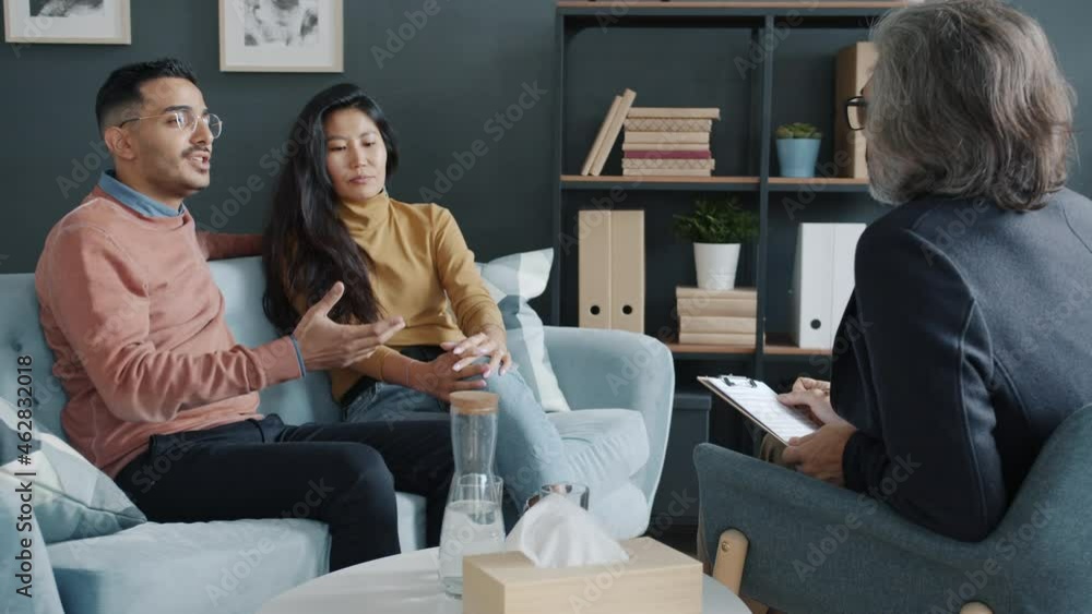 Middle Eastern guy and Asian girl are fighting during family therapy session with psychologist while mature man specialist is listening. Relationship and problems concept. Middle Eastern guy and Asian girl are fighting during family therapy session with psychologist while mature man specialist is listening. Relationship and problems concept.
