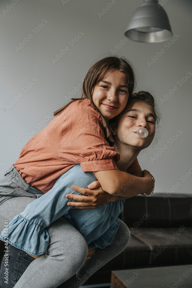 Playful girl blowing bubble gum while piggybacking friend against wall at home Playful girl blowing bubble gum while piggybacking friend against wall at home