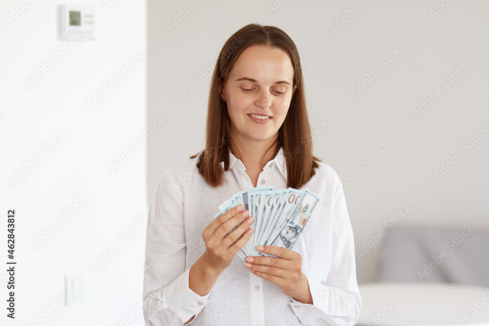 Smiling rich attractive woman with dark hair wearing white casual style shirt, counting money while posing at home in light room, having satisfied facial expression. Smiling rich attractive woman with dark hair wearing white casual style shirt, counting money while posing at home in light room, having satisfied facial expression.