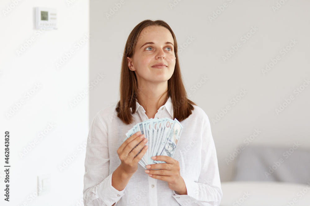 Portrait of attractive woman with dark hair wearing white casual style shirt, looking away with dreamy and satisfied facial expression, dreaming about excellent purchase. Portrait of attractive woman with dark hair wearing white casual style shirt, looking away with dreamy and satisfied facial expression, dreaming about excellent purchase.