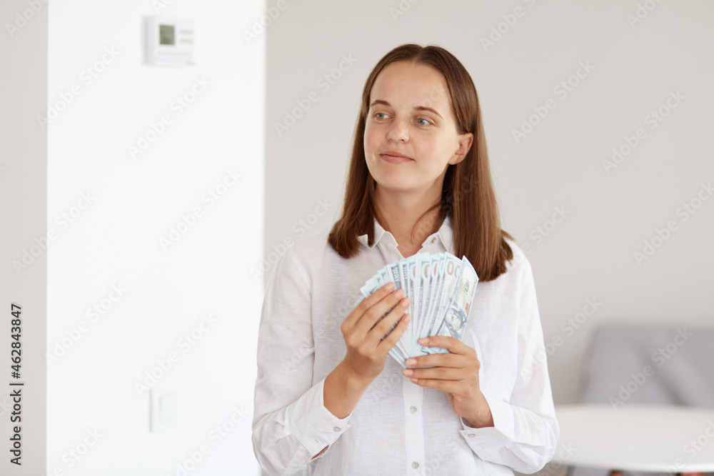 Smiling attractive woman with dark hair wearing white casual style shirt, holding fan of money in hands, looking away, planing to buy expensive purchase. Smiling attractive woman with dark hair wearing white casual style shirt, holding fan of money in hands, looking away, planing to buy expensive purchase.