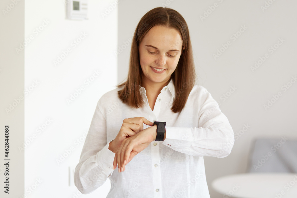 Portrait of good looking young adult woman wearing white casual style shirt, using her wrist watch, looking at smart phone screen, posing indoor in light room. Portrait of good looking young adult woman wearing white casual style shirt, using her wrist watch, looking at smart phone screen, posing indoor in light room.