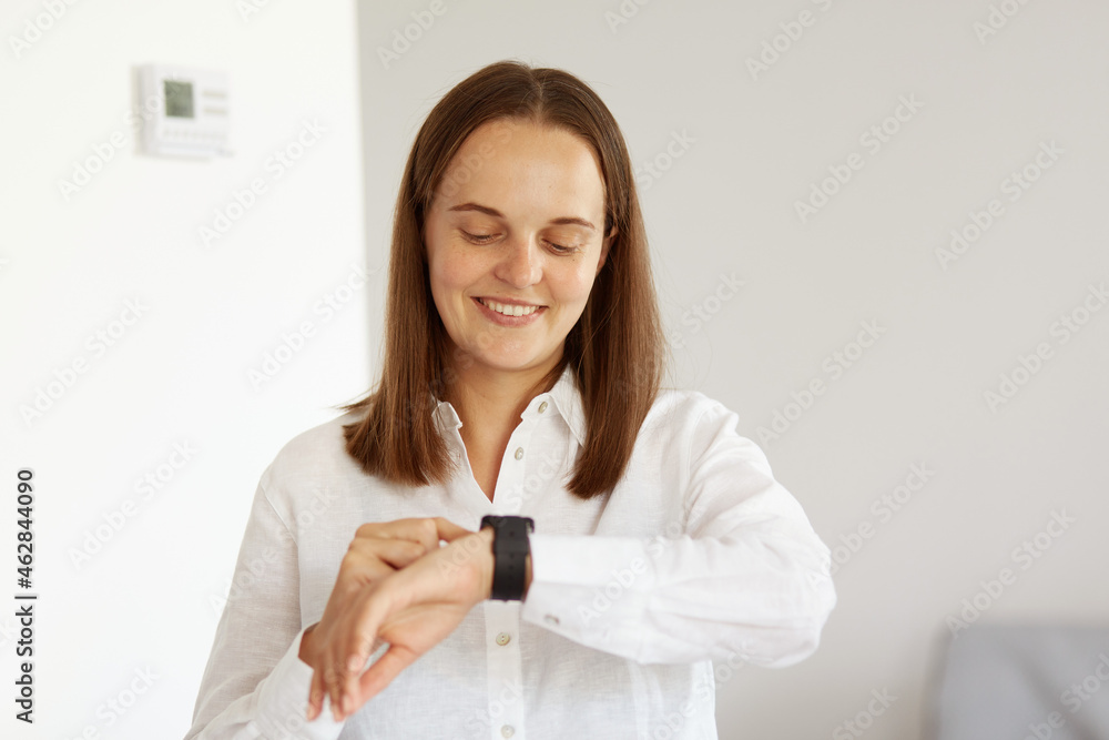 Smiling young adult female with pleasant appearance wearing white casual style shirt standing using smartwatch, looking and touching touchscreen, expressing happiness, modern technology. Smiling young adult female with pleasant appearance wearing white casual style shirt standing using smartwatch, looking and touching touchscreen, expressing happiness, modern technology.