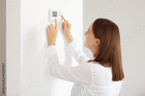 Obraz na plátně Good looking young adult woman wearing white casual style shirt, using connected appliances device on wall, monitoring energy security heating system, digital control in apartment