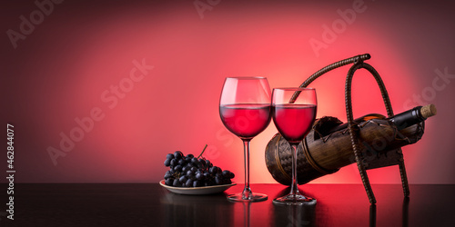 Two wine glasses with red wine and bottle of wine on the table on red background with copy space. Banner. © Sergey
