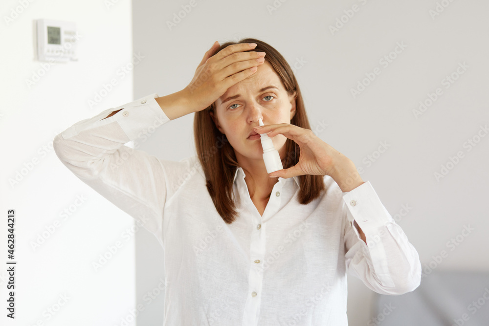 Unhealthy winsome female with pleasant appearance wearing white casual style shirt, using nasal spray for runny nose, feels flu, catching cold, suffering headache. Unhealthy winsome female with pleasant appearance wearing white casual style shirt, using nasal spray for runny nose, feels flu, catching cold, suffering headache.