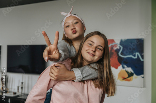 Smiling girl piggybacking friend showing victory sign while standing at home photo