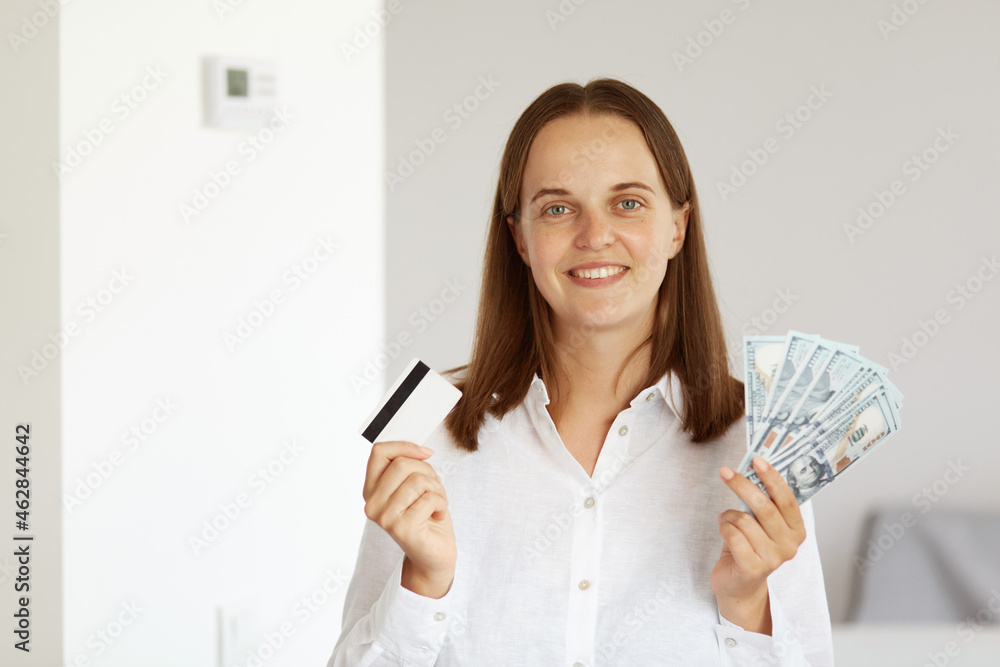 Satisfied rich young adult woman wearing white casual style shirt, posing in light room at home, showing to camera credit card and big fan of dollar banknotes, banking. Satisfied rich young adult woman wearing white casual style shirt, posing in light room at home, showing to camera credit card and big fan of dollar banknotes, banking.