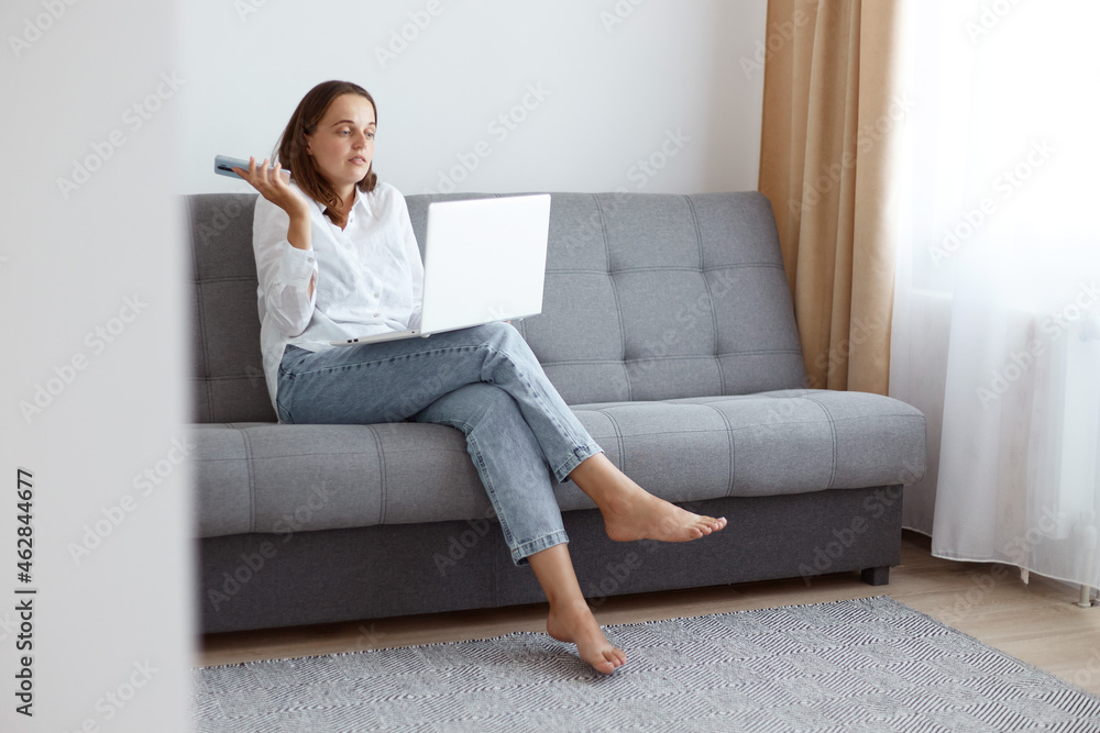 Indoor shot of puzzled dark haired female wearing white casual style shirt and jeans, sitting on sofa in living room, holding cell phone in hands, having helpless confused expression. Indoor shot of puzzled dark haired female wearing white casual style shirt and jeans, sitting on sofa in living room, holding cell phone in hands, having helpless confused expression.