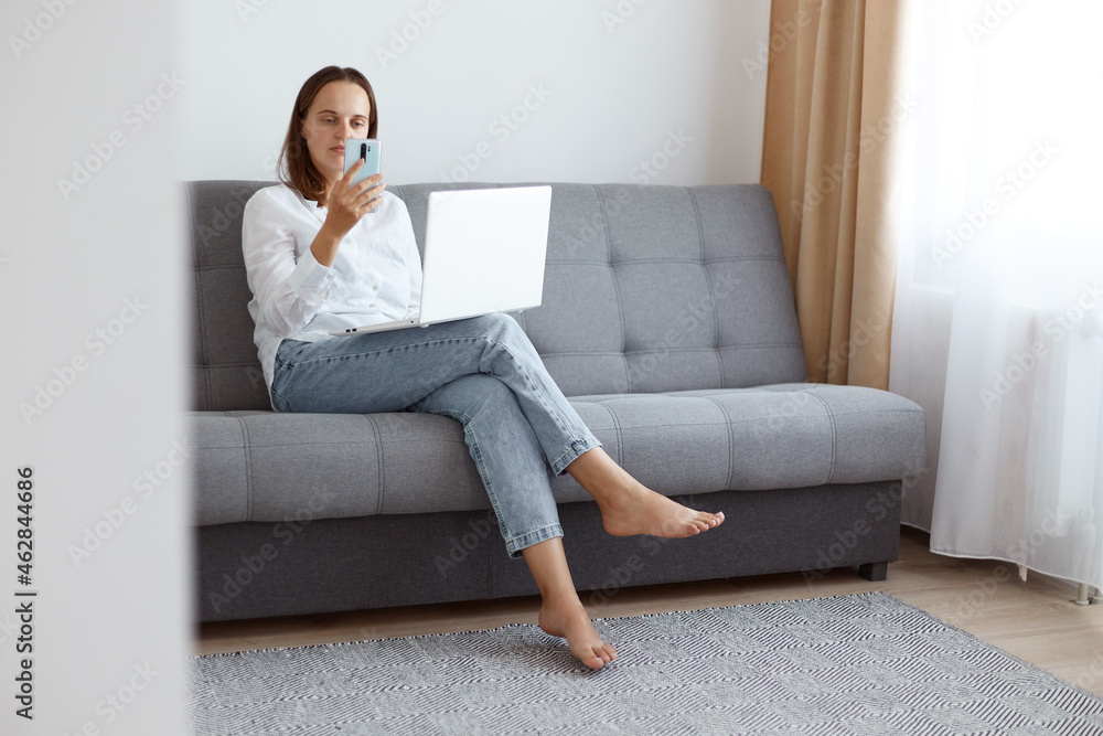 Attractive young adult female freelancer, wearing white casual style shirt and jeans, sitting on gray couch with laptop, using smart phone, looking at device screen. Attractive young adult female freelancer, wearing white casual style shirt and jeans, sitting on gray couch with laptop, using smart phone, looking at device screen.