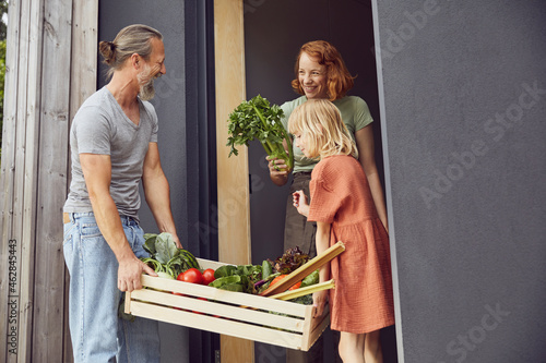 Smiling woman holding vegetable while father and daughter carrying crate at entrance