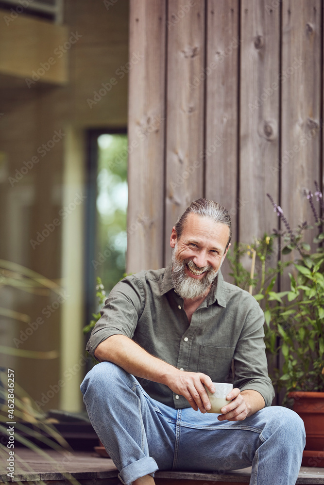 Cheerful mature man holding coffee cup while sitting outside tiny house Cheerful mature man holding coffee cup while sitting outside tiny house
