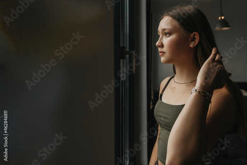Thoughtful teenage girl looking through window while standing at home photo