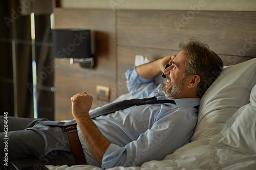 Smiling businessman lying on the bed in hotel room talking on the phone photo