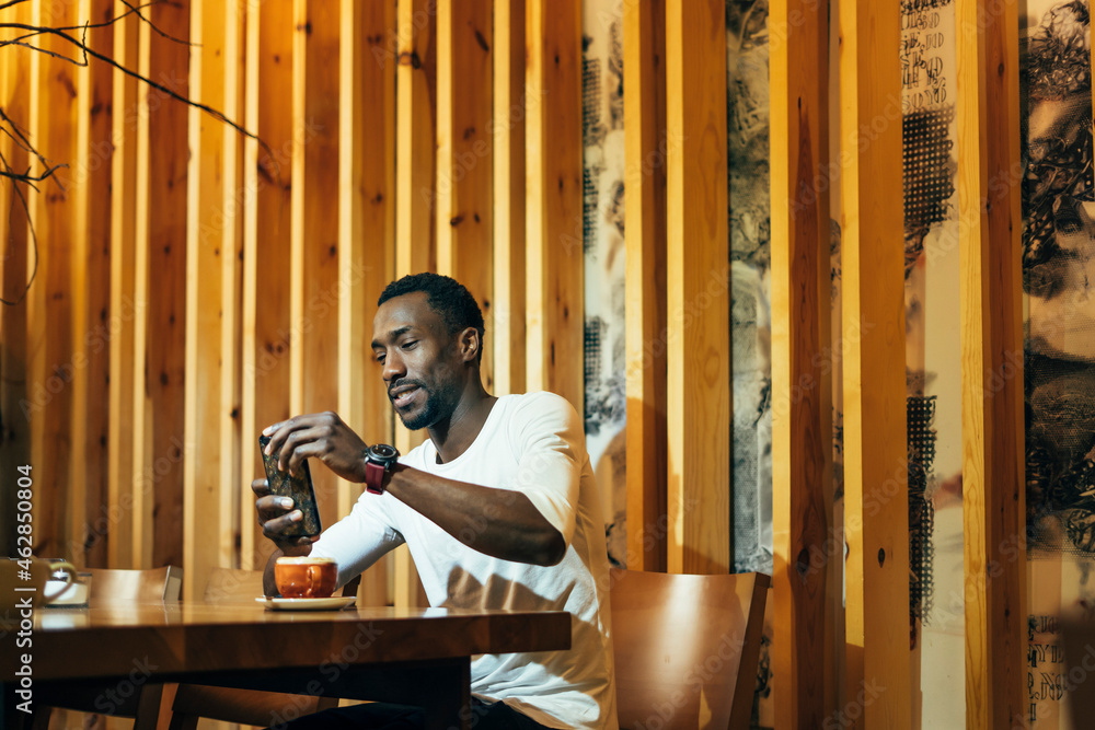 Smiling young man using smart phone while sitting with coffee on table against wall in cafe Smiling young man using smart phone while sitting with coffee on table against wall in cafe