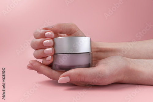 Nice female hands with natural manicure holding a glass cosmetics jar on soft pink background.