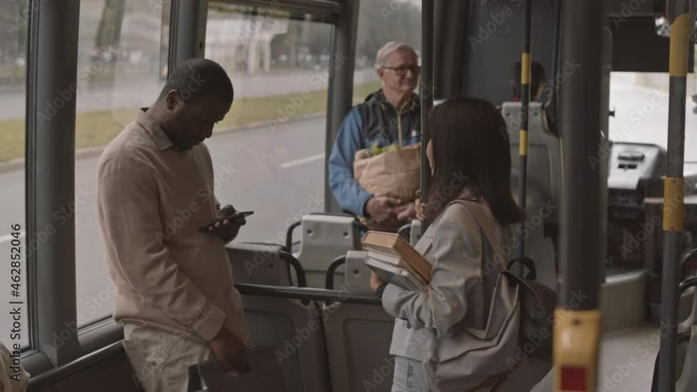 Medium long of young Black man using smartphone and his Asian female companion who holding books, standing in public transport, talking Medium long of young Black man using smartphone and his Asian female companion who holding books, standing in public transport, talking