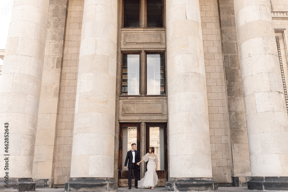 Young couple, man and woman, bride and groom are standing on large concrete steps among columns holding hands, looking at each other. Marriage concept of happy family life. Young couple, man and woman, bride and groom are standing on large concrete steps among columns holding hands, looking at each other. Marriage concept of happy family life.
