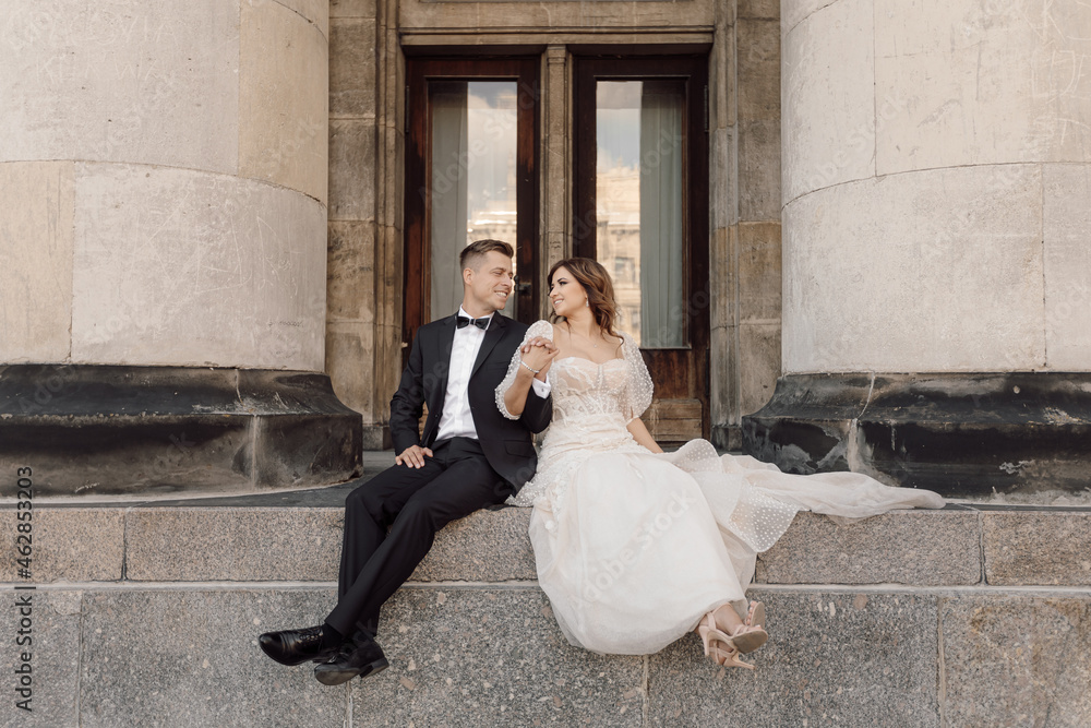 Young couple, man and woman, bride and groom are sitting on large concrete steps among columns holding hands, looking at each other. Marriage concept of happy family life.