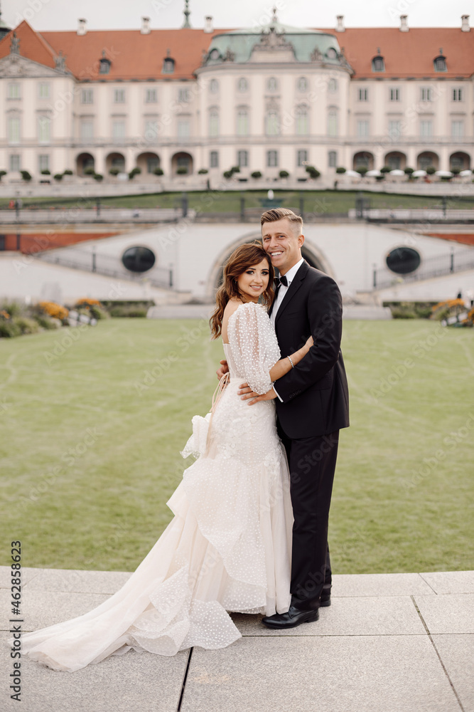Naklejka premium photo of bride and groom in full-length wedding dress standing in embrace against background of European old town, smiling and looking at camera.