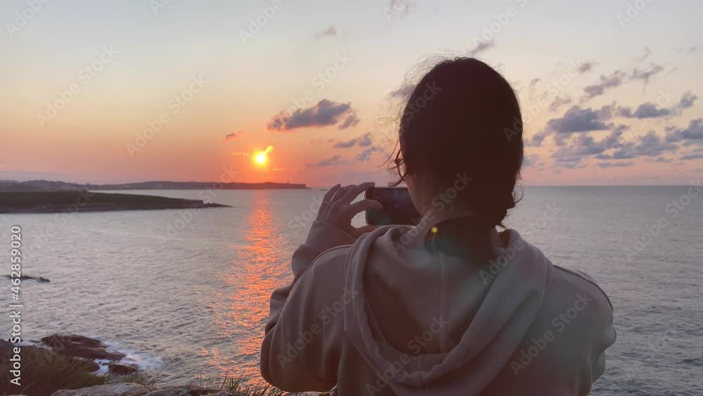 A teenager takes photos with her mobile of the sunset at the edge of the sea A teenager takes photos with her mobile of the sunset at the edge of the sea