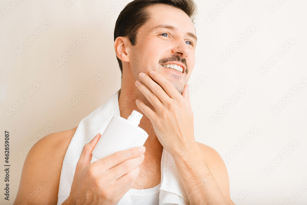 Charming happy young man with towel on shoulders applying moisturizing after shave lotion, handsome guy touching cheek with fine bristles and holding bottle of cosmetic gel. Male skin care and beauty Charming happy young man with towel on shoulders applying moisturizing after shave lotion, handsome guy touching cheek with fine bristles and holding bottle of cosmetic gel. Male skin care and beauty