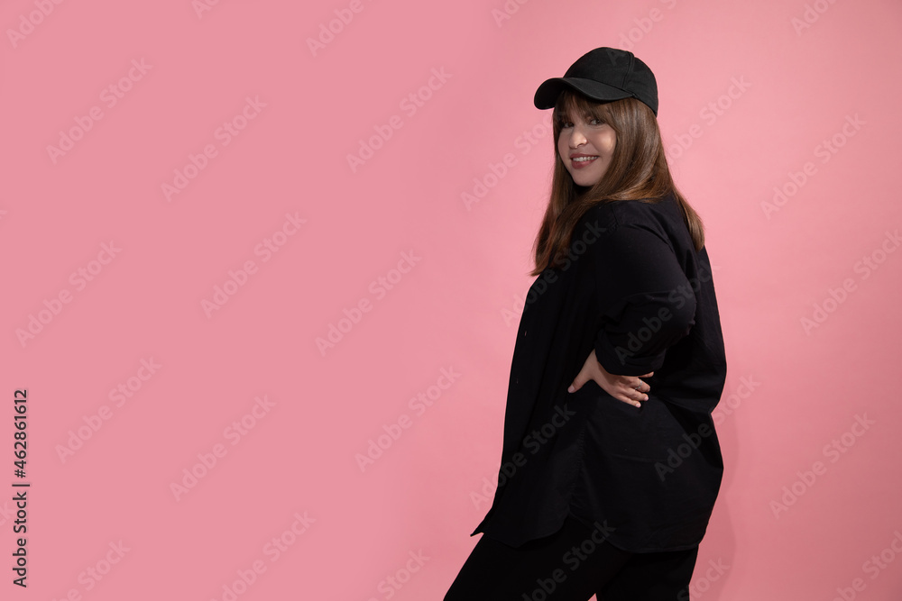A young beautiful woman in black casual clothes and sports baseball cap stands with back half-sided to the camera and looks into frame on pink studio background. A young beautiful woman in black casual clothes and sports baseball cap stands with back half-sided to the camera and looks into frame on pink studio background.