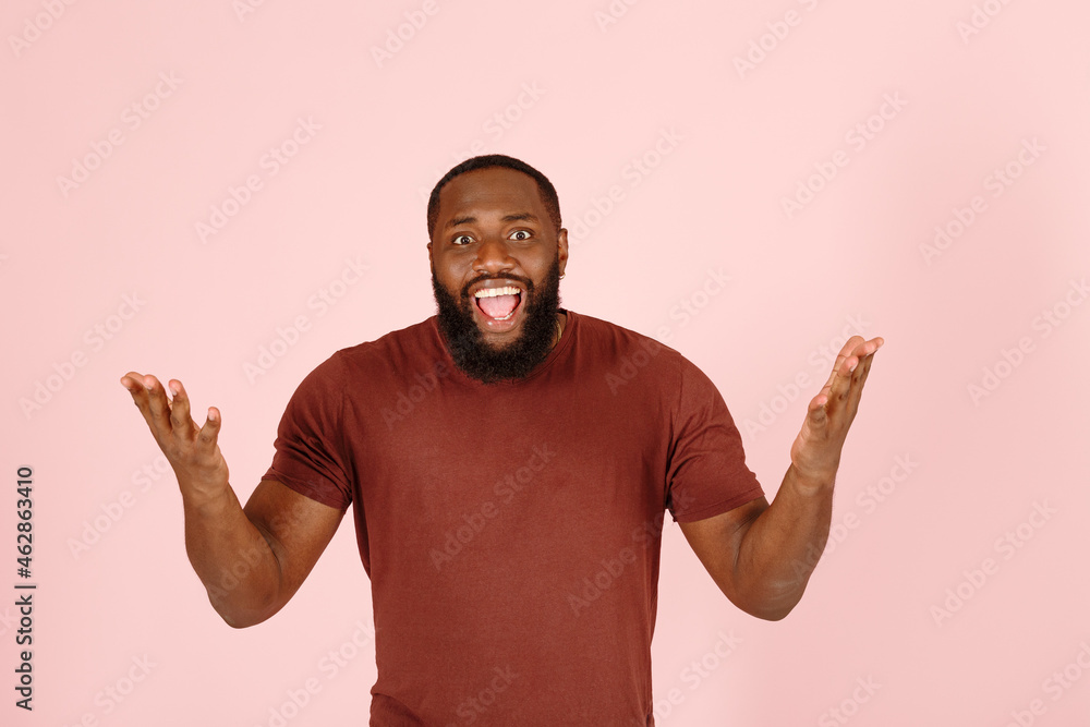 Joyful broadbrow young African-American man actor in casual brown t-shirt poses for camera on light pink background in studio Joyful broadbrow young African-American man actor in casual brown t-shirt poses for camera on light pink background in studio