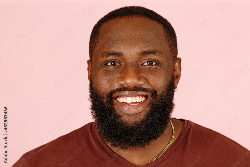 Handsome bearded African-American model man in brown t-shirt smiles to camera on pink background in studio extreme close view Handsome bearded African-American model man in brown t-shirt smiles to camera on pink background in studio extreme close view