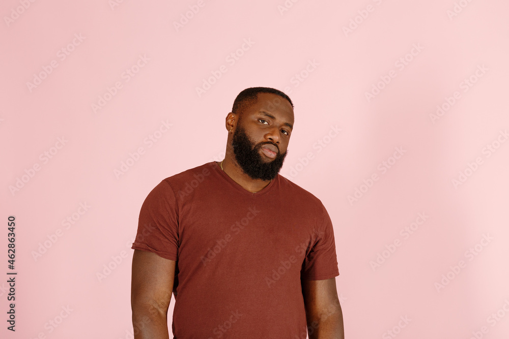 Skeptical bearded African-American man in brown t-shirt poses for camera on pink background in studio close view Skeptical bearded African-American man in brown t-shirt poses for camera on pink background in studio close view