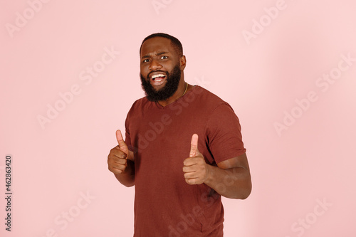 Confused bearded black male actor in brown t-shirt shows thumbs-up posing for camera on pink background in studio close view