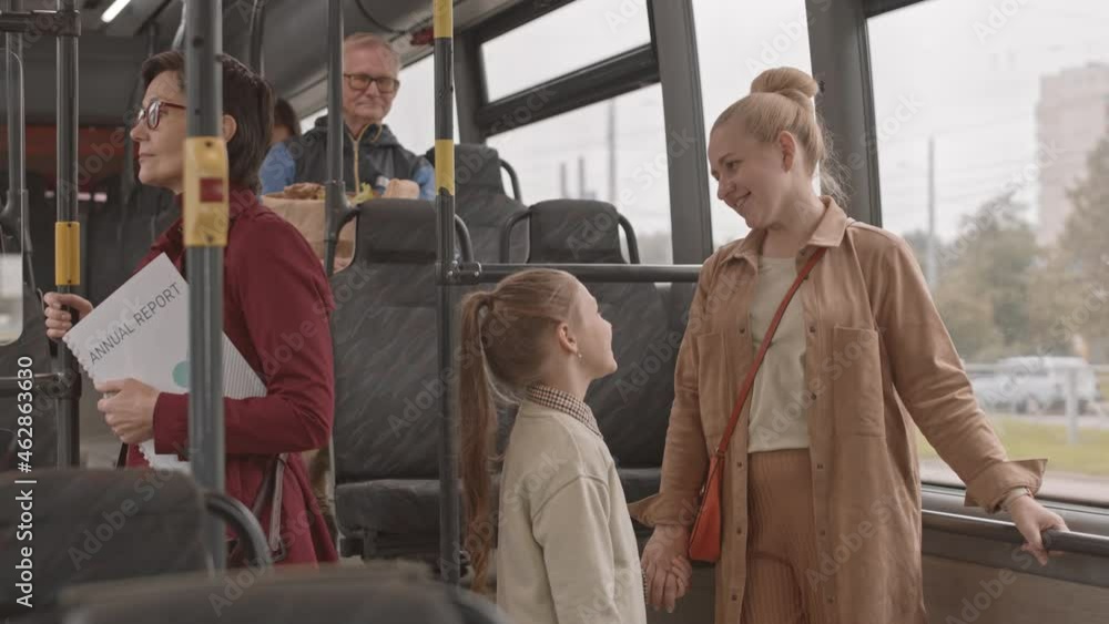 Medium long of blonde Caucasian woman holding hands with talking to little daughter, standing by window on bus at daytime Medium long of blonde Caucasian woman holding hands with talking to little daughter, standing by window on bus at daytime