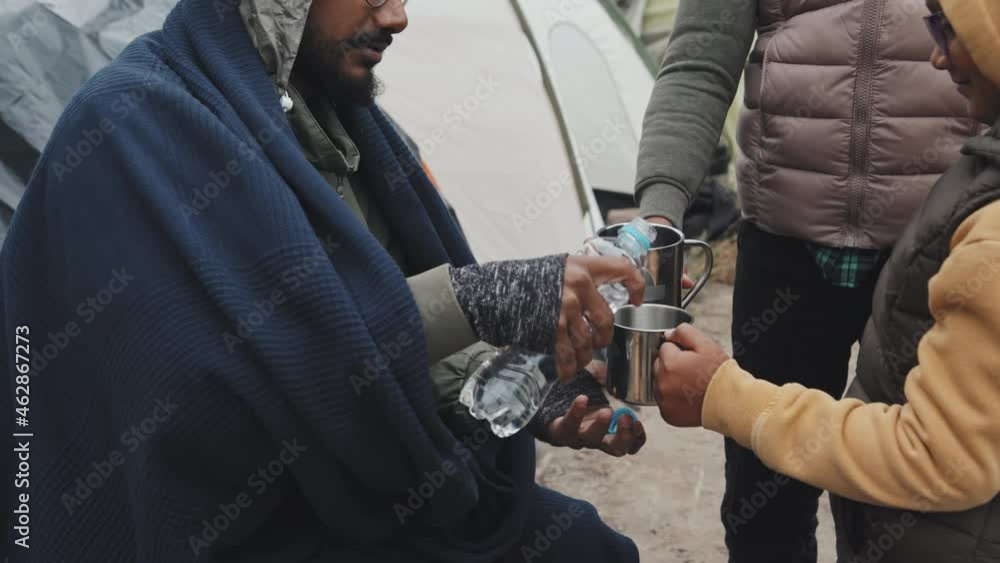 Handheld shot of Arab man pouring little amount of water in metal cups for refugee children. Residents of tent city in lack of water Handheld shot of Arab man pouring little amount of water in metal cups for refugee children. Residents of tent city in lack of water