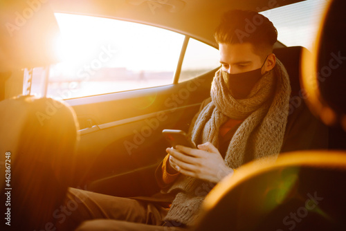 Young man in protective sterile mask in taxi car on a back seat  checking her cell phone. Safety and pandemic concept. Work on the way to the office. Covid-2019.