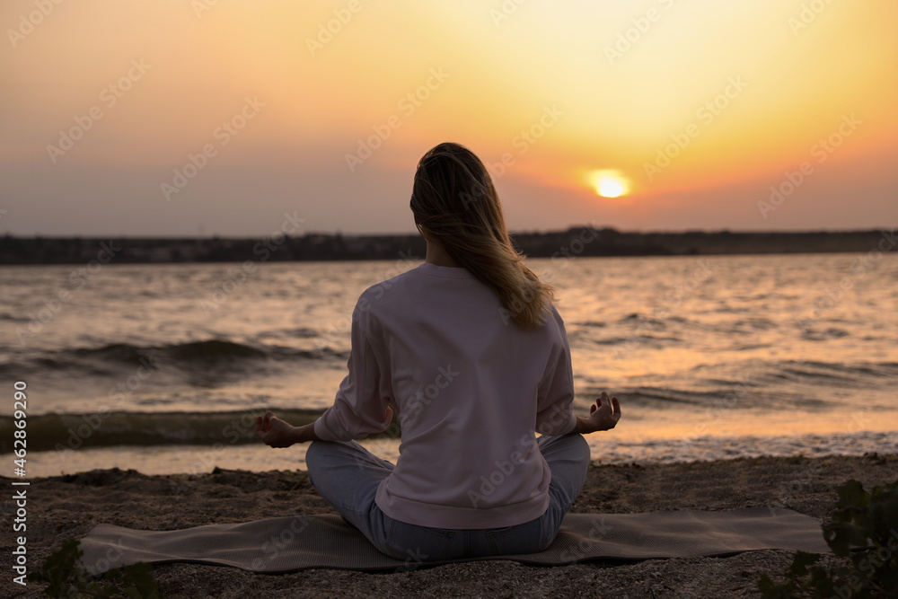 Young woman meditating on beach near river at sunset, back view Young woman meditating on beach near river at sunset, back view